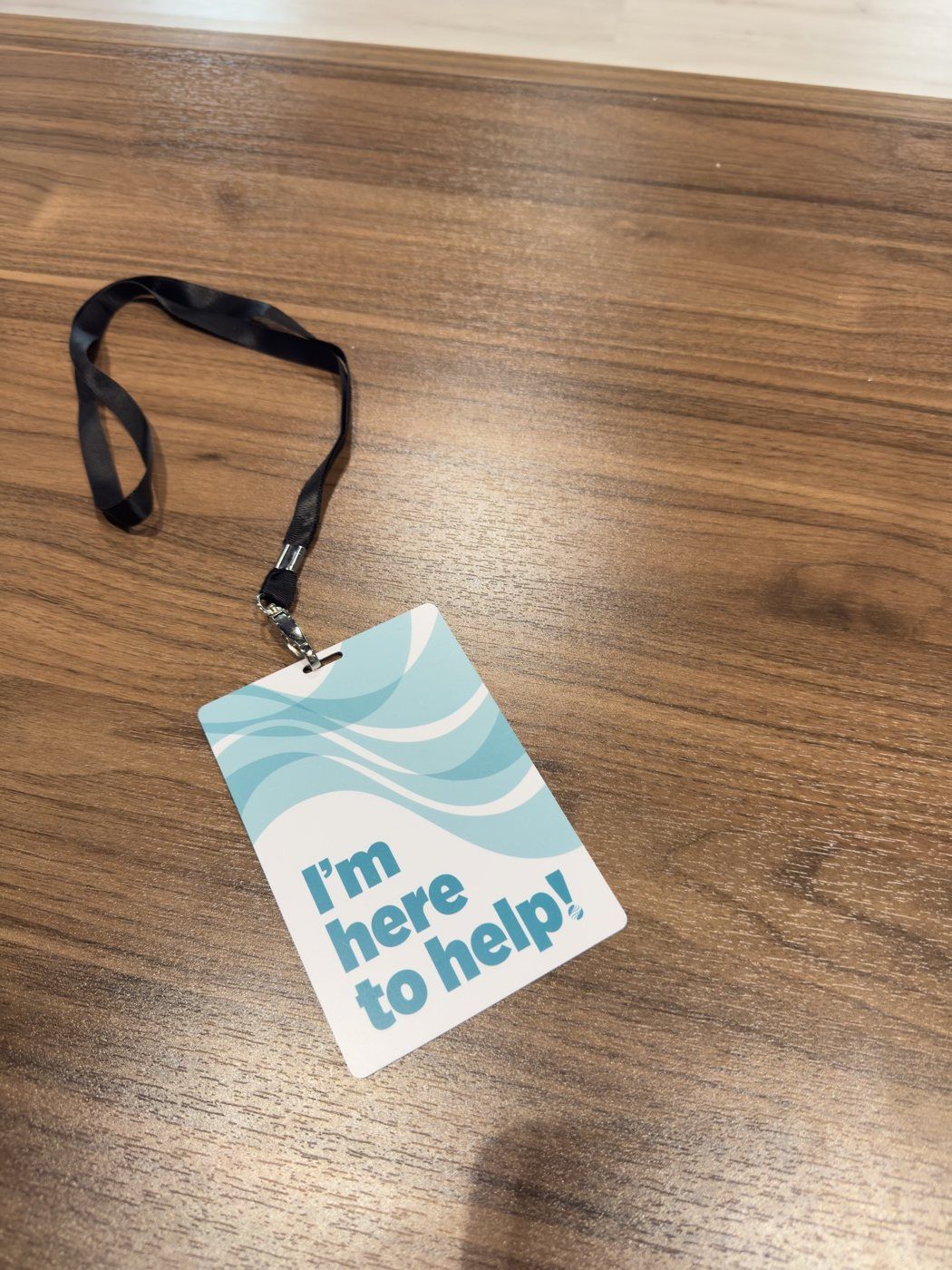 Printed lanyard card reading I'm here to help, in layered blue waves, photographed on a wood table.